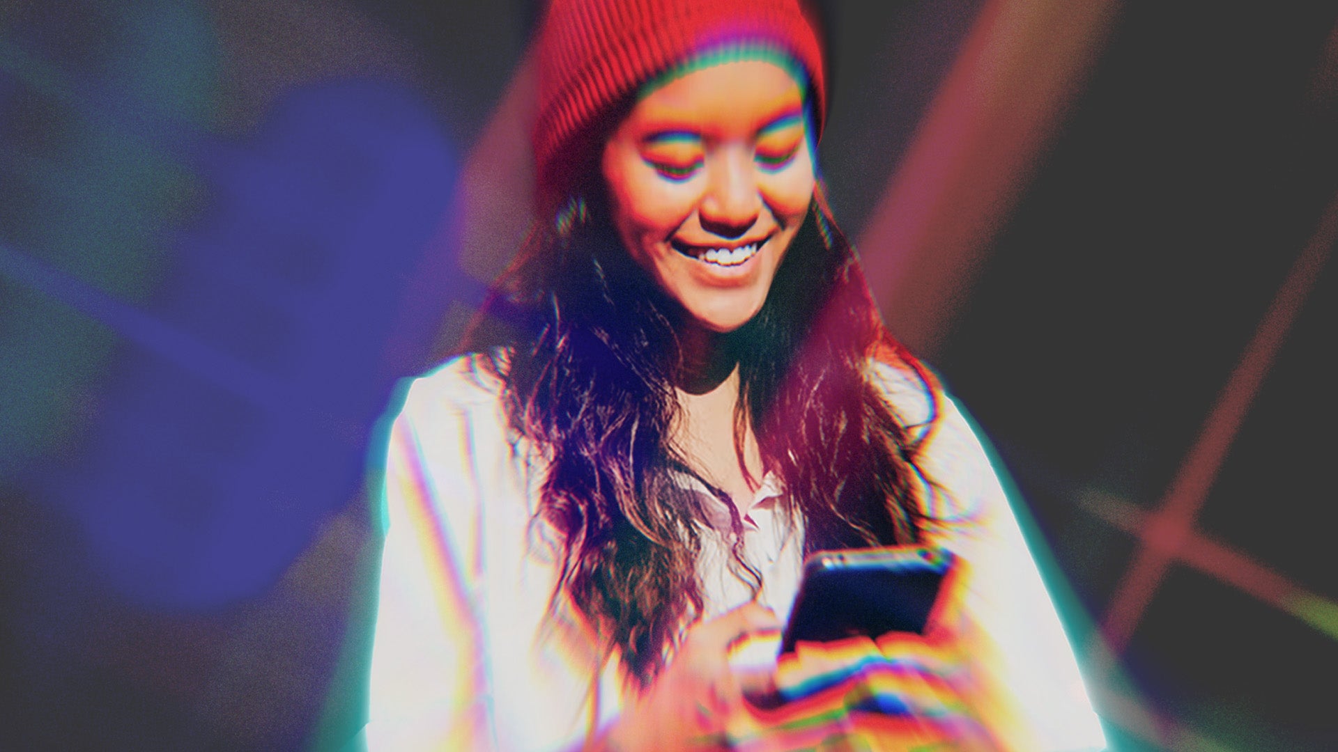 A smiling woman looking at a phone wearing a red hat and white top. She is standing against a dark background